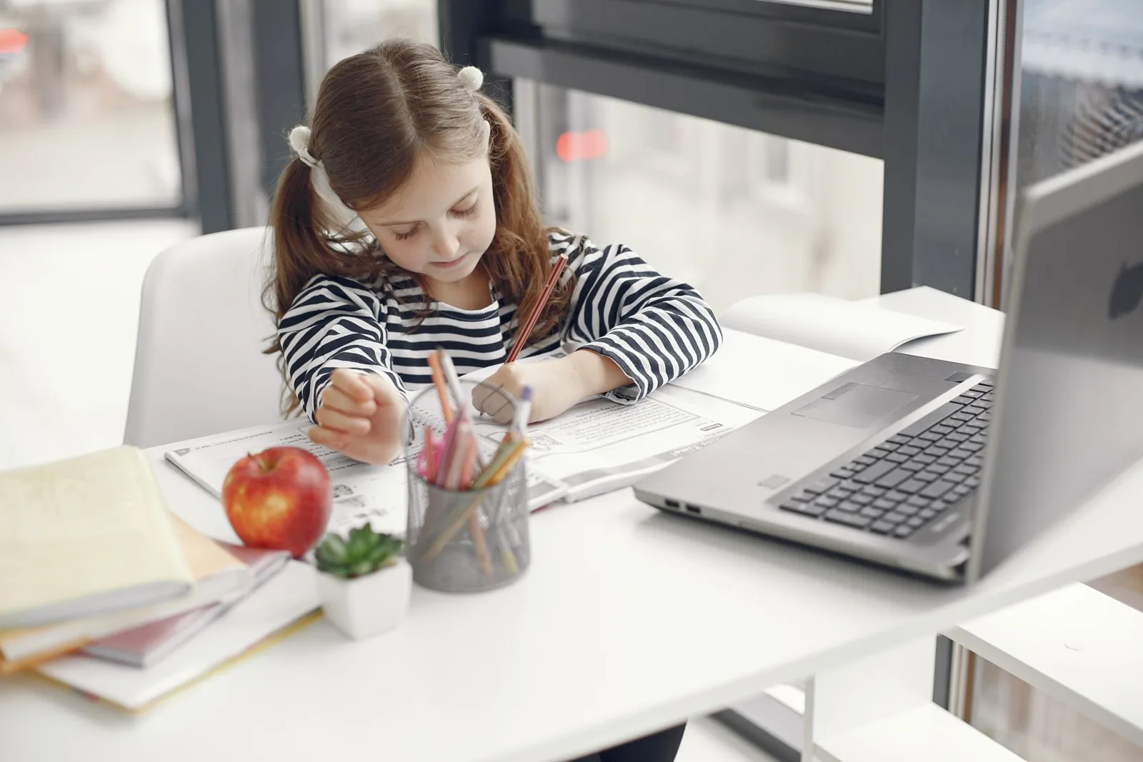 Niña escribe en su cuaderno en un escritorio ordenado, junto a un computador
