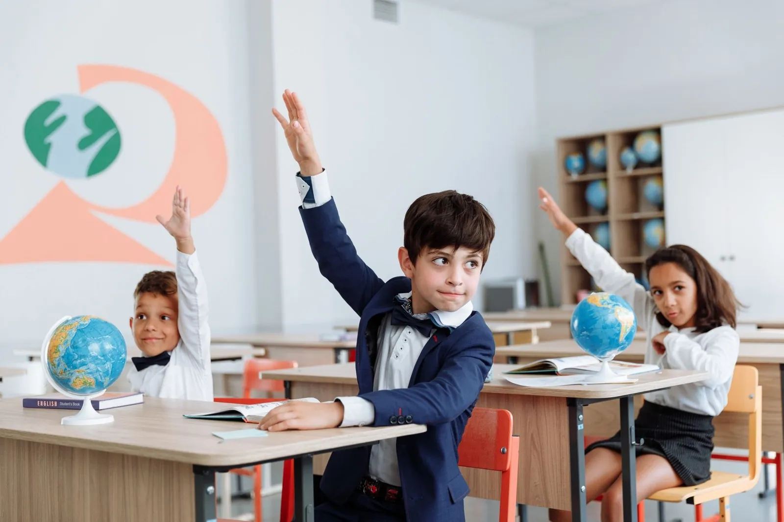 Niños con uniforme levantan la mano en una sala con globos terráqueos y un libro de inglés sobre la mesa