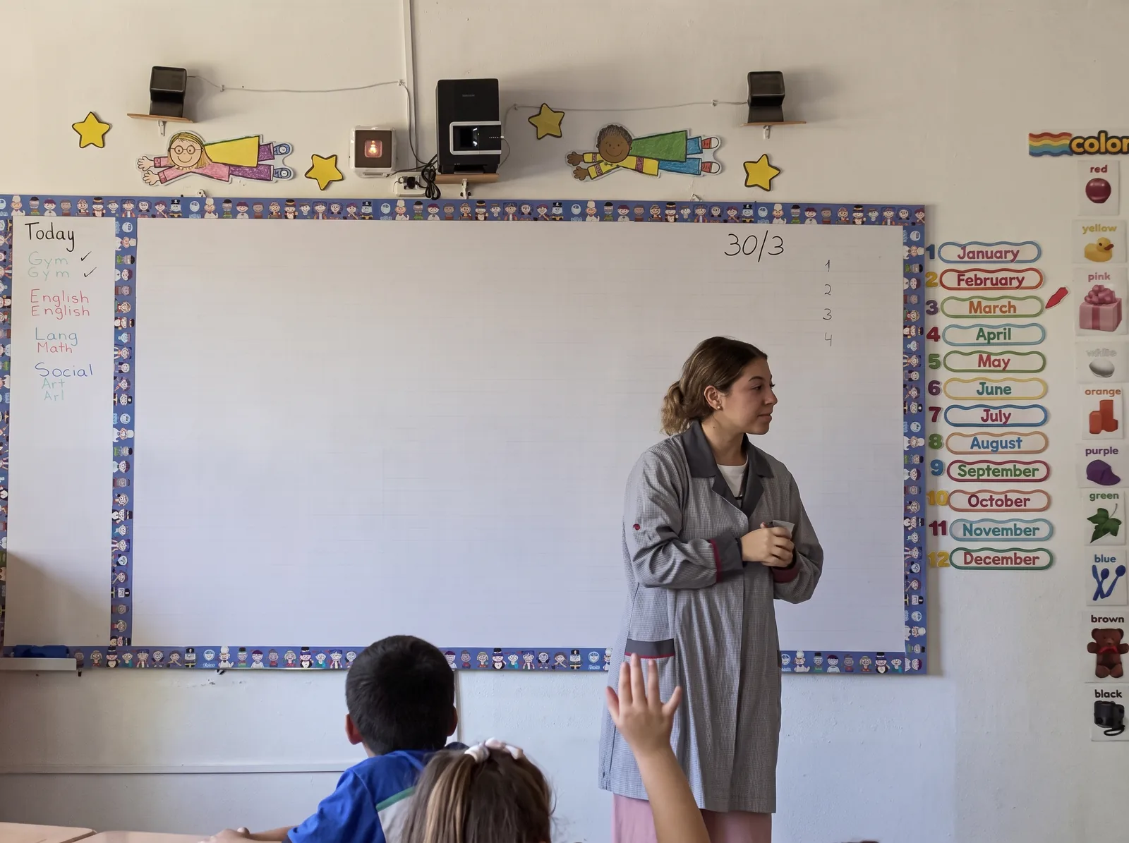 Isidora Benito frente a la pizarra de una sala de básica, junto a la lista de los meses en inglés, mientras una alumna levanta la mano