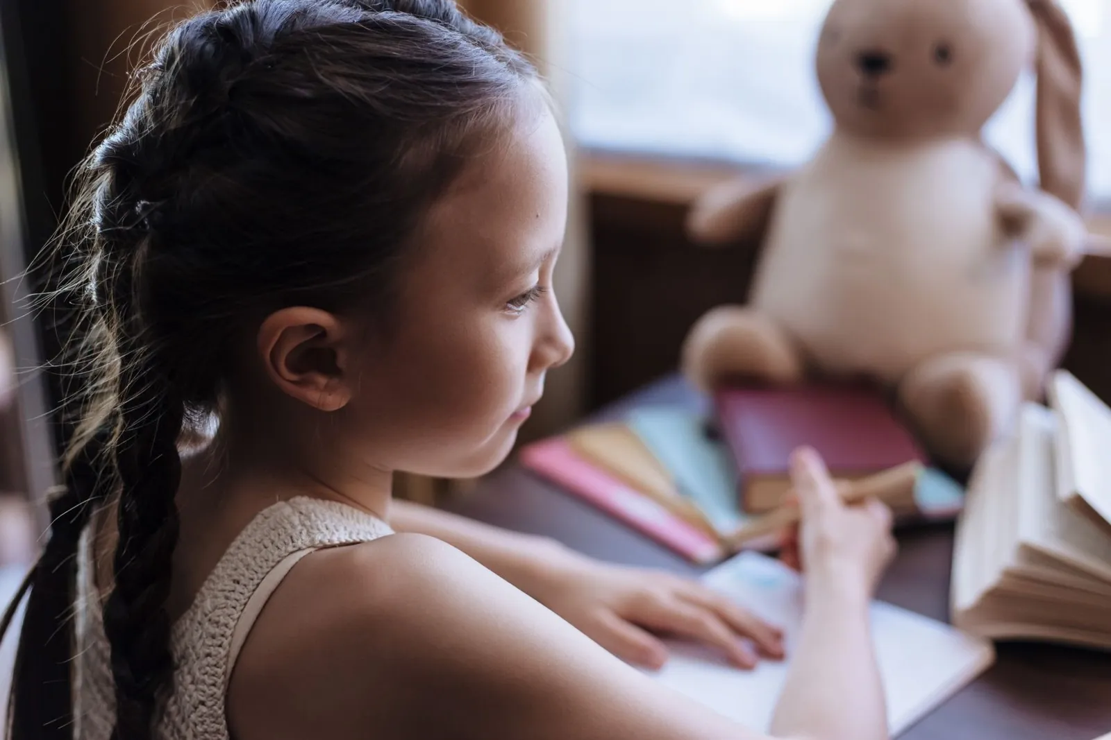 Niña con trenzas escribe en su cuaderno, con un oso de peluche y libros de colores a su lado